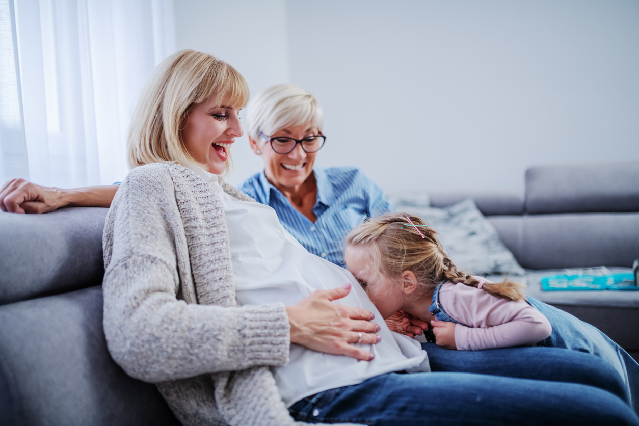 A woman in her mid-30s celebrates being pregnant through fertility treatment as her young daughter kisses her belly and grandma watches with a smile.
