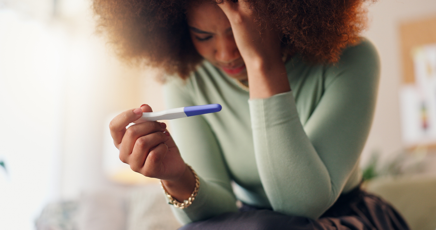 A young woman holds a negative pregnancy test in her hand and hangs her head in frustration and sadness after a failed IVF cycle.