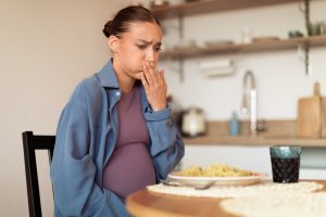 Pregnant woman sits at her kitchen table, showing signs of nausea because of the food in front of her.